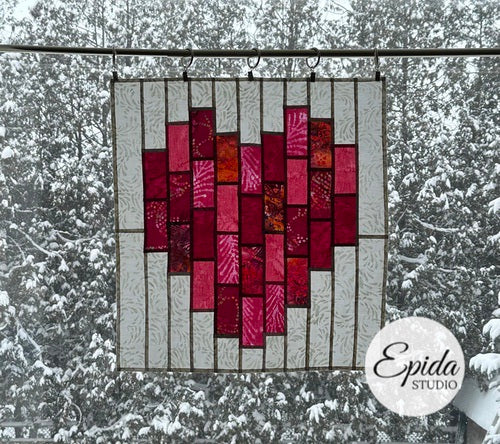 Stained glass heart-shaped panel with red and pink colors hanging outdoors against a snowy background.