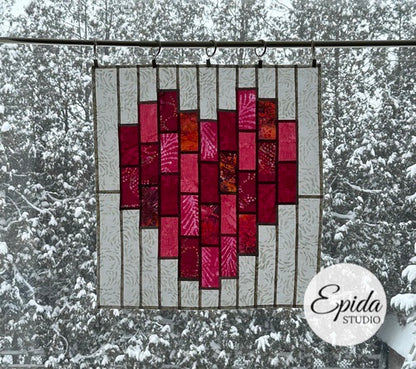 Stained glass heart-shaped panel with red and pink colors hanging outdoors against a snowy background.
