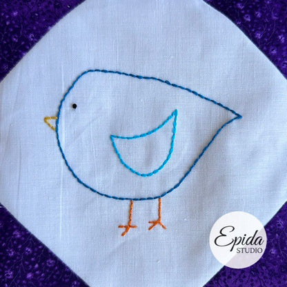 Embroidered bird design on a white fabric square with a purple border, featuring the brand 'Epida Studio'.