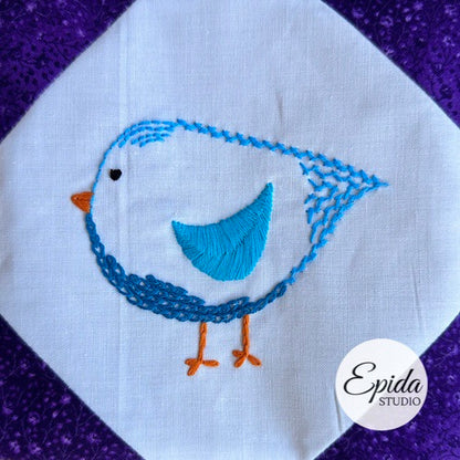 Embroidered blue bird design on white fabric with a purple background, featuring the Epida Studio logo.