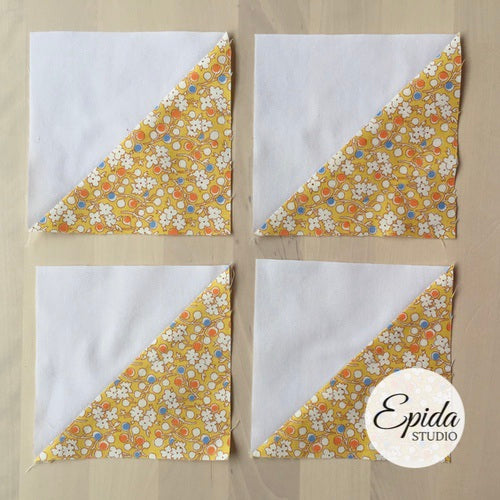 Four fabric blocks arranged in a 2x2 grid on a light wood surface. Each block is split diagonally, with one half in white fabric and the other half in a yellow floral print fabric with white, orange, and blue flowers.