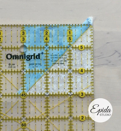 A clear Omnigrid quilting ruler with yellow markings and a blue-and-white patterned paper sheet placed on top, photographed on a light marble-like surface. An Epida Studio watermark appears in the lower right corner.