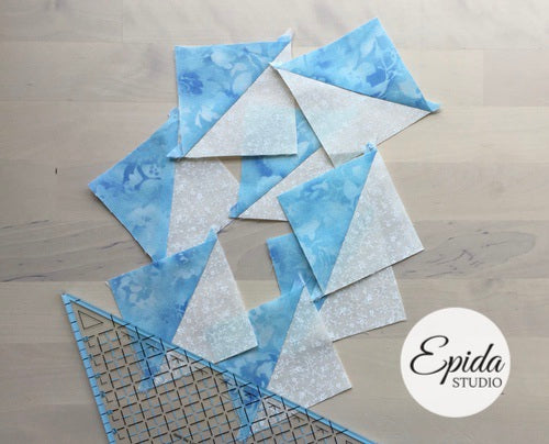 Quilted fabric triangles in blue floral and white textured patterns arranged on a light wood floor, with a quilting ruler and a white circular 'Epida Studio' logo overlay.