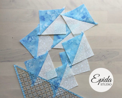 Quilted fabric triangles in blue floral and white textured patterns arranged on a light wood floor, with a quilting ruler and a white circular 'Epida Studio' logo overlay.